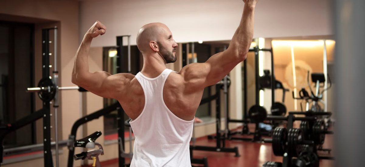 A muscular man in a gym flexes his back muscles wearing a white tank top.