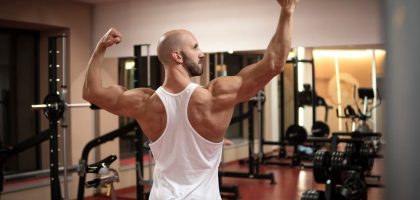 A muscular man in a gym flexes his back muscles wearing a white tank top.