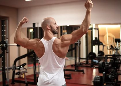 A muscular man in a gym flexes his back muscles wearing a white tank top.